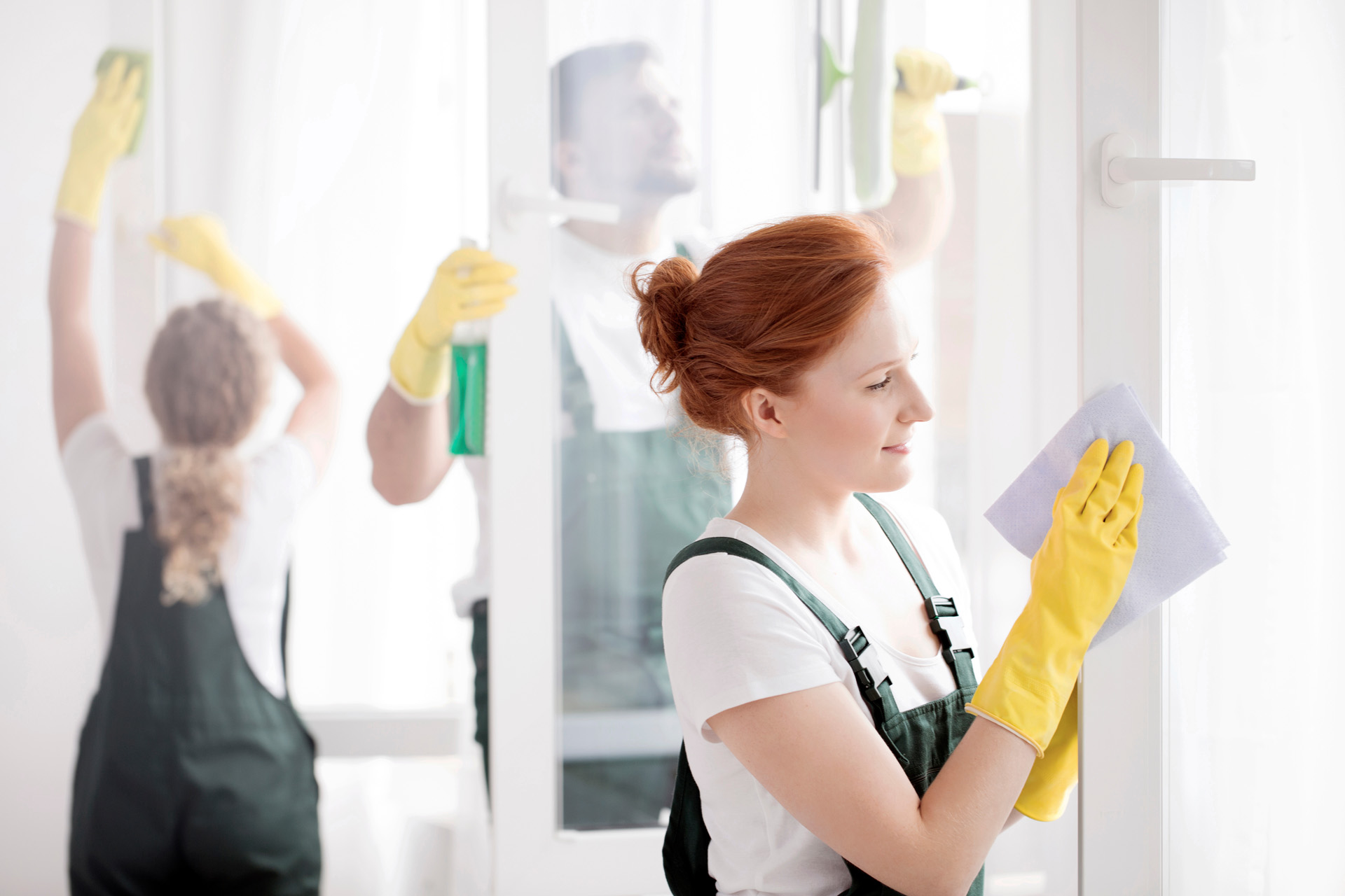 A ginger woman and her two coworkers in yellow gloves and green overalls cleaning windows